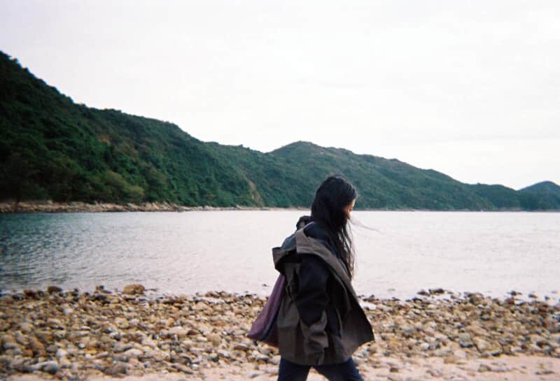 A person walking along a rocky beach with mountains in the background and a cloudy sky.