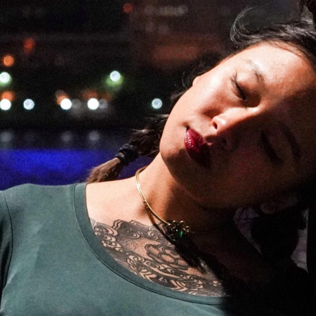 A close-up portrait of Tim Tim Cheng with braided hair, wearing a green top and a gold choker. The background features softly blurred city lights, creating a warm evening atmosphere.